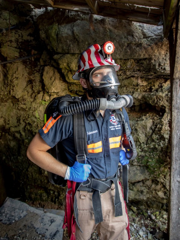 A student standing at the entrance to a mine wearing a mining uniform a backpack a hardhat with a head lamp blue latex type gloves a gas mask with tubes leading to the backpack with a face shield built into the gas mask